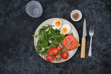 A plate with a salmon sandwiche, arugula, tomatoes, boiled egg with glass of water on black background. Healthy food. Horizontal frame.