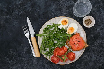 A plate with a salmon sandwiche, arugula, tomatoes, boiled egg with glass of water on black background. Healthy food. Horizontal frame.