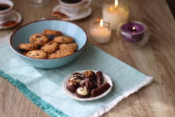 Plate of chocolate pralines, bowl of cookies, cups of tea, glasses of juice and lit candles on the table. Selective focus.