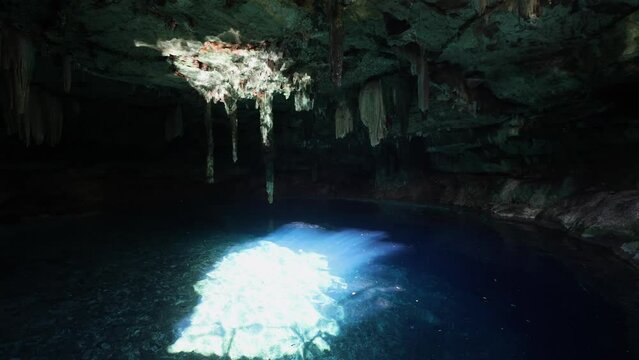 panning shot of subterranean pool with crystal blue waters and a bright sun spot in the middle with light reflections on the roof with stalactites