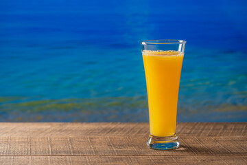 Freshly squeezed orange juice in glass on wooden table with sea water background on sunny summer day in tropical beach cafe, close up, copy space. Glass of fresh fruit orange juice