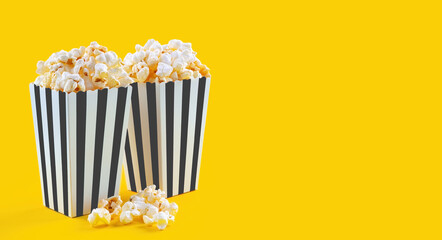 Two black white striped carton buckets with tasty cheese popcorn, isolated on yellow background. Box with scattering of popcorn grains. Fast food, movies, cinema and entertainment concept.