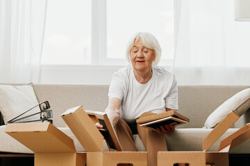 elderly woman sits on a sofa at home with boxes. collecting things with memories albums with photos and photo frames moving to a new place cleaning things and a happy smile. Lifestyle retirement.