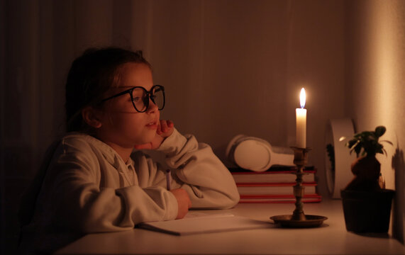 Schoolgirl uses candlelight to writes and reads in darkness without electric lights at home. Teen with poor eyesight wears glasses to studies and to do school homework during blackout. Vision problems