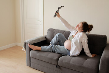 Happy expecting mother and popular vlogger is recording video for online blog sits on sofa in...
