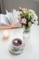 a spring still life, a bouquet of roses in a vase, a cup of tea and an book on white table. spring greeting card with spring flowers. Romantic background