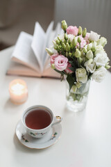 a spring still life, a bouquet of roses in a vase, a cup of tea and an book on white table. spring greeting card with spring flowers. Romantic background
