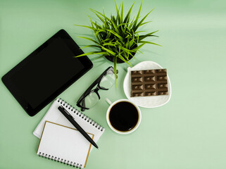 Top view office desk with tablet, stationery and coffee cup and chocolate bar.  Flat workspace with sunlight and copy space, green background.  The concept of online learning, webinars, office work.