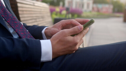 Obraz na płótnie Canvas Close up young busy businessman sits on bench with purple flowers in flower beds behind him and use mobile phone. man in formal suit and tie is texting on smartphone, looking for information, trader