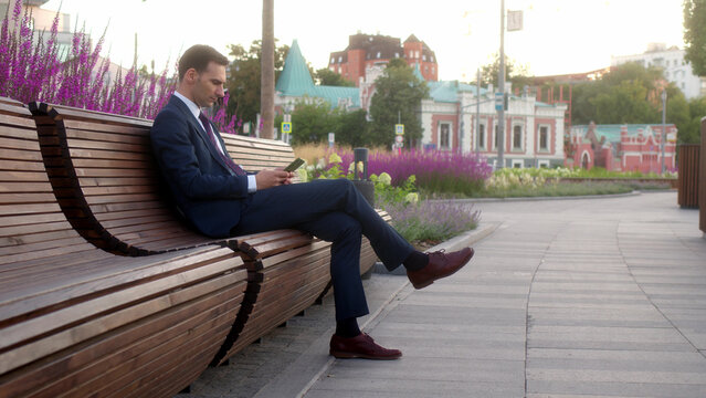 Young businessman in official dark blue suit sits on bench in park before meeting with partners and preparing to make presentation. Office clerk at lunch break working on cell or smartphone outdoors.