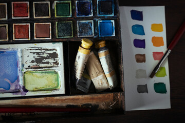 A vintage metal watercolor painting set with pans and tubes of paint against a dark wooden desk