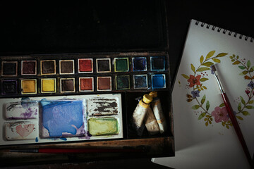 Vintage watercolor painting set with a painting of flowers on a spiral bound pad on a dark wood desk 