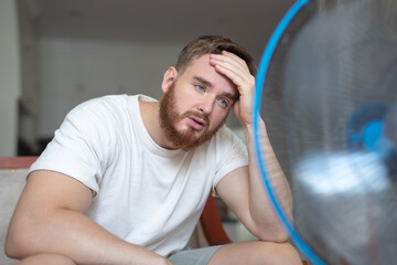 Young bearded man using electric fan at home, sitting on couch cooling off during hot weather,...