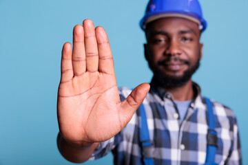 Serious looking construction worker in coveralls doing hand stop in dissapproval on blue background. Professional builder worker doing denial sign having displeased attitude.