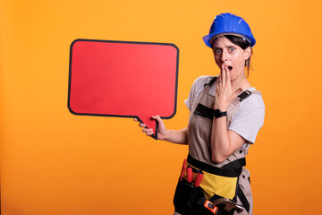 Young renovating supervizor holding empty cardboard banner for advertisement, using carton copyspace. Woman builder showing red speech bubble with mockup on yellow background in studio.