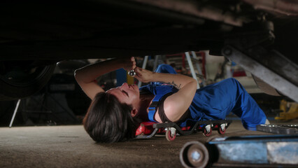 woman mechanic with tattoos lies under car on car repair trolley and repairs it with tools. View of...