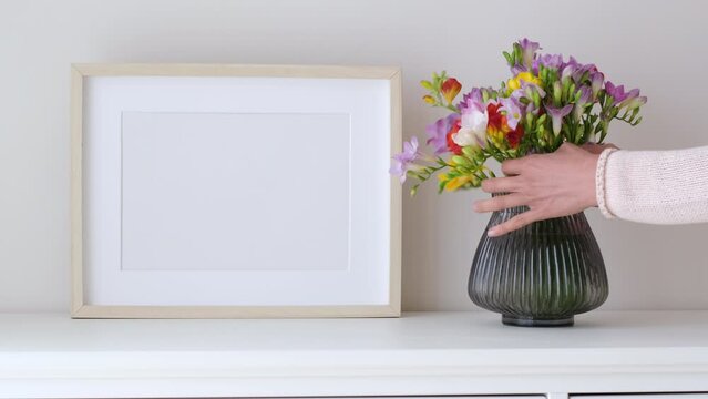 Female hands puts flowers in a vase. Wooden frame mockup in white room interior, empty mock-up with copy space. Light frame with a vase in the interior for design presentation.
