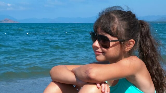 Child in swimsuit at sunny weather. A pretty little girl in sunglasses sits on the beach and enjoy the sunny sea weather against blue bay.