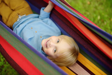 Fototapeta na wymiar Cute little blond white boy enjoy and having fun with multicolored hammock in backyard or outdoor playground. Summer outdoors active leisure for kids. Child