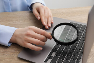 Woman holding magnifier near laptop at wooden table, closeup. Online searching concept