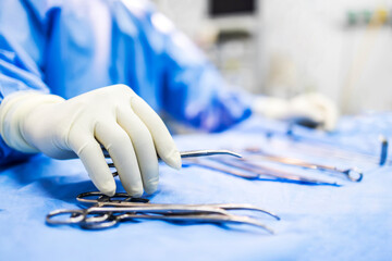 Hand of doctor or surgeon in blue gown pick up surgical clamp instrument or tools inside operating theatre with blur background.Medical equipment for surgery inside room in hospital.Modern technology. - obrazy, fototapety, plakaty