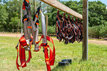 A wooden rack with multiple white, black and orange colored rock climbing equipment, cords, harnesses, hooks, fasteners, cams, belts, slings and cables hanging from a board outside in a grassy field. 