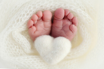 Knitted white heart in the legs of a baby. Macro photography. The tiny foot of a newborn baby. Soft feet of a new born in a white wool blanket. Close up of toes, heels and feet of a newborn.