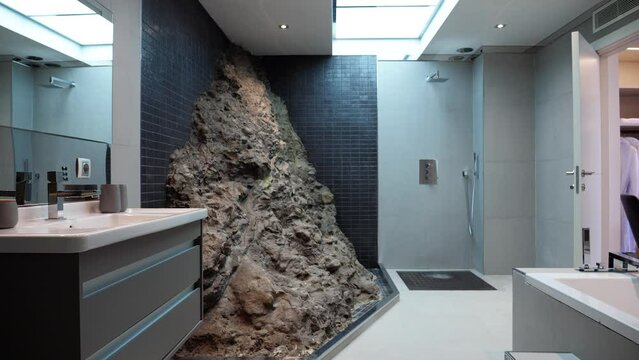 a modern and impressive bathroom with a rock wall in the center, a modern sink with mirror on the left and a bathtub on the right, a shower at the back, a skylight on the ceiling