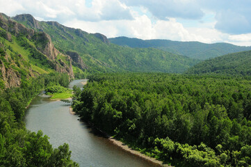 A wide river flowing through dense taiga at the foot of a high mountain range under a summer cloudy sky.