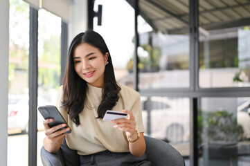 Attractive Asian female holding her smartphone and credit card, using mobile banking app