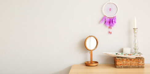 Stylish dream catcher hanging on light wall and table with mirror, candles and basket. Banner for design