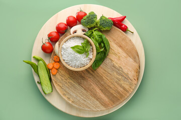 Wooden cutting board with bowl of sea salt and fresh vegetables on color background