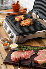 Wooden board with raw meat and modern electric grill on table