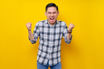 angry young Asian man in a plaid shirt screaming and clenching first isolated on yellow background. people lifestyle concept