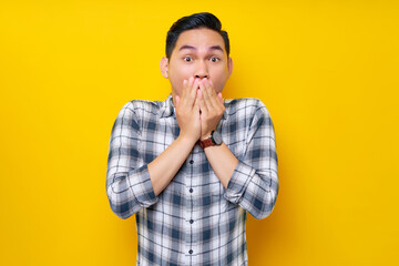 Handsome young Asian man surprised in plaid shirt covers his mouth with his hand isolated on yellow...