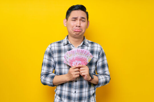 Portrait of crying young Asian man wearing plaid shirt holding cash money in rupiah banknotes and thrilled to get money isolated on yellow background. people lifestyle concept