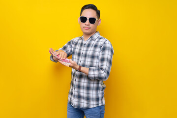 Portrait of smiling young Asian man wearing plaid shirt and sunglasses holding cash money in rupiah banknotes, Rich people show arrogance isolated on yellow background. people lifestyle concept - obrazy, fototapety, plakaty