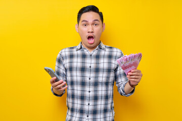 Portrait of Surprised young Asian man wearing plaid shirt holding mobile phone and cash money in rupiah banknotes isolated on yellow background. people lifestyle concept