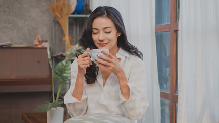 Young Asian woman, freelance, artist or student enjoy drinking a coffee in the morning with cozy, warm, freedom, relaxing atmosphere. Concept of weekend break time, inspiration time, wellness daytime.