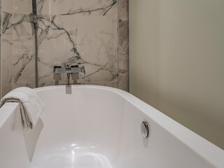 White acrylic bathtub. Close-up. The towel is thrown over the edge. On the beige marble tile wall...