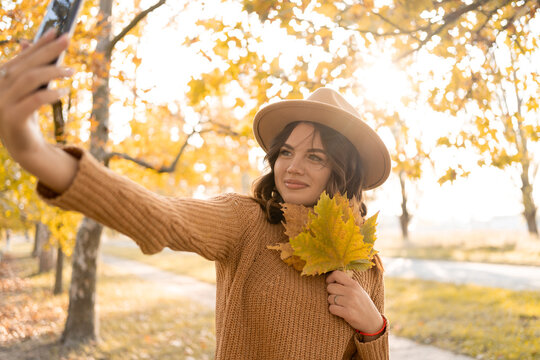 Beautiful young Caucasian woman with fallen leaves taking a selfie with smartphone outdoors in park in autumn. Magnificent brunette girl taking picture of herself while walking
