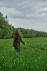 Obraz premium a woman in a long raincoat runs across a field in tall green grass in cloudy weather in spring