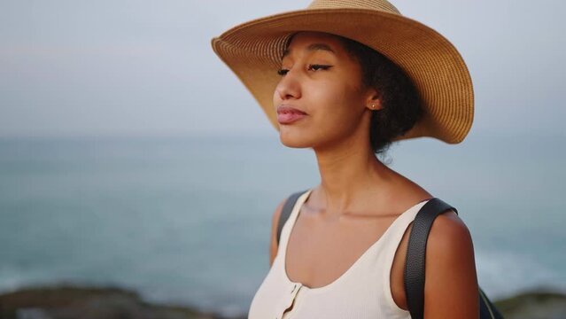 Pretty african woman enjoys ocean view standing on island on sunrise. Biracial female in straw hat sightseeing on travel to exotic sea destination. Tourist bipoc girl relax. Tracking shot.