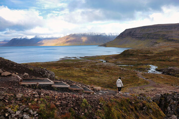 A lone traveler walks through the rugged Icelandic landscape towards a serene fjord, surrounded by...