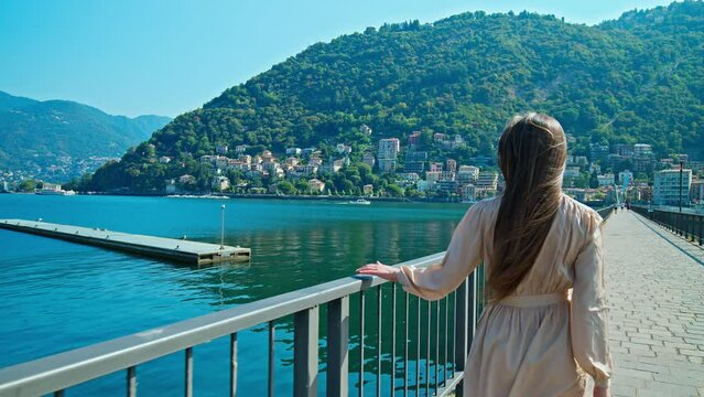 Beautiful Girl with long hair in a dress walking in slow motion by Lake Como. A female tourist walks by the port of Como and enjoys the panoramic view of the lake with boats in Italy in summer.