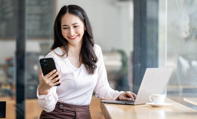 Attractive asian woman smiling and looking at mobile phone in her hand, sitting at counter table in cafe with laptop computer.