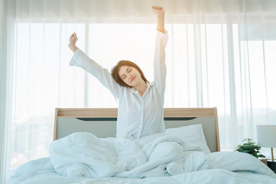 Photo of young happy woman in pajama stretching her arms and smiling while sitting on bed after sleep