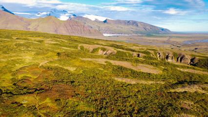 Stunning Icelandic landscape featuring dramatic volcanic mountains, vast moss-covered fields, and a powerful waterfall under a clear blue sky. Perfect for showcasing Iceland’s raw natural beauty and u