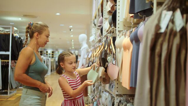 Mother and daughter choose looking for clothes in shopping mall. Young mother and child chooses clothes in sports clothing boutique. Cheerful child and mother choosing sports clothes in retail store