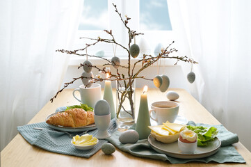 Breakfast table with hanging Easter eggs on spring branches, candles, and blue gray decoration in front of a window with white curtains, copy space, selected focus